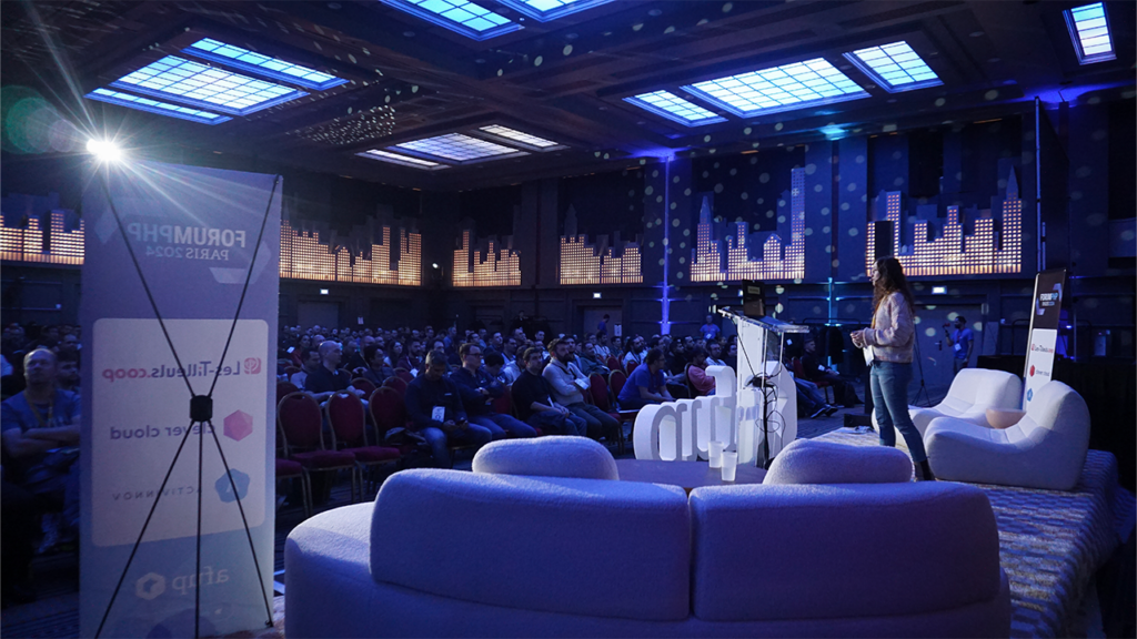Une conférencière est sur scène, la salle baigne d'une ambiance bleutée, lors du Forum PHP 2024